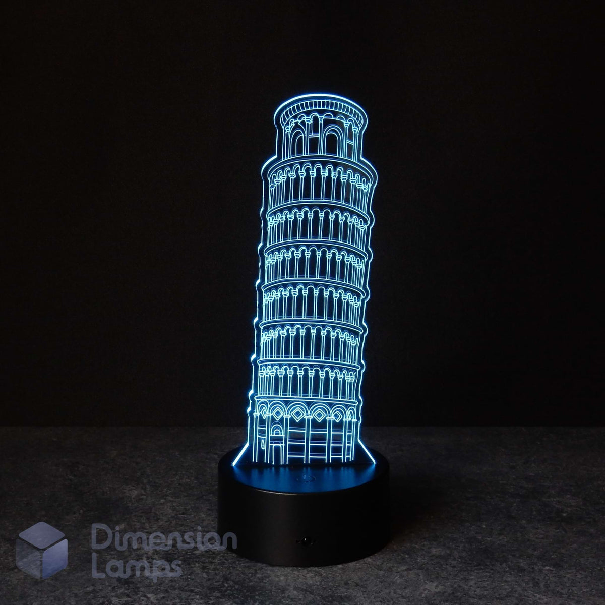 A 3D LED night light shaped like the Leaning Tower of Pisa, glowing with a bright blue outline on a black circular base against a dark background. A 'Dimension Lamps' watermark is visible in the bottom-left corner.