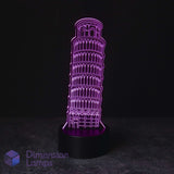 A 3D LED acrylic lamp featuring the Leaning Tower of Pisa, glowing with a vibrant purple light from a black base on a dark gray surface against a black background. A 'Dimension Lamps' logo is visible in the bottom-left corner.