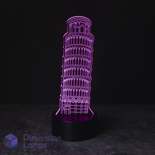 A 3D LED acrylic lamp featuring the Leaning Tower of Pisa, glowing with a vibrant purple light from a black base on a dark gray surface against a black background. A 'Dimension Lamps' logo is visible in the bottom-left corner.