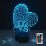 A heart-shaped 3D optical illusion night light with the word 'LOVE' in 3D block letters, shown glowing with a blue cyan light on a dark surface. A 16 colour remote control is shown. Heart-shaped 'LOVE' lamp is a perfect girls gift idea.