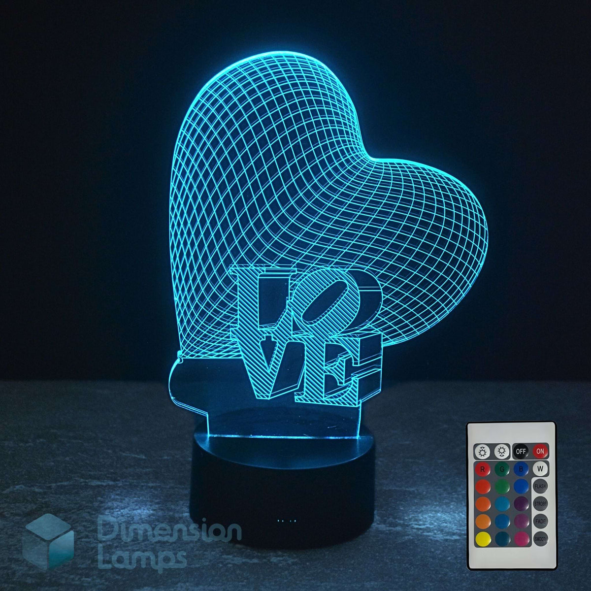 A heart-shaped 3D optical illusion night light with the word 'LOVE' in 3D block letters, shown glowing with a blue cyan light on a dark surface. A 16 colour remote control is shown. Heart-shaped 'LOVE' lamp is a perfect girls gift idea.