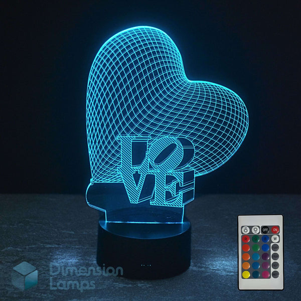 A heart-shaped 3D optical illusion night light with the word 'LOVE' in 3D block letters, shown glowing with a blue cyan light on a dark surface. A 16 colour remote control is shown. Heart-shaped 'LOVE' lamp is a perfect girls gift idea.