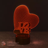 A heart-shaped 3D optical illusion lamp with a wireframe heart design and the word 'LOVE' integrated into the base, illuminated in a warm orange-red glow on a dark surface against a dark background. The Love Heart 3D LED lamp, radiating a warm glow, is a perfect gift for girls across Australia.