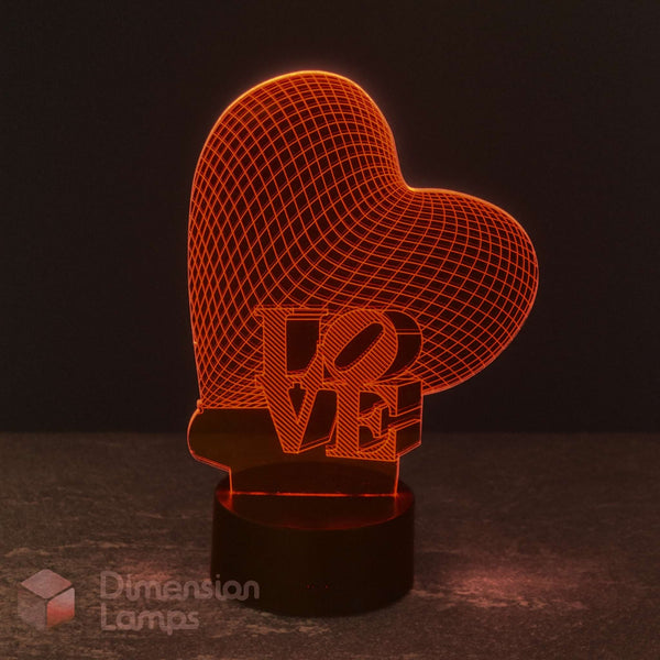 A heart-shaped 3D optical illusion lamp with a wireframe heart design and the word 'LOVE' integrated into the base, illuminated in a warm orange-red glow on a dark surface against a dark background. The Love Heart 3D LED lamp, radiating a warm glow, is a perfect gift for girls across Australia.