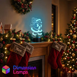 A glowing Santa Claus 3D LED lamp on a fireplace mantel surrounded by Christmas decorations. The 'Dimension Lamps' logo in the bottom left corner.