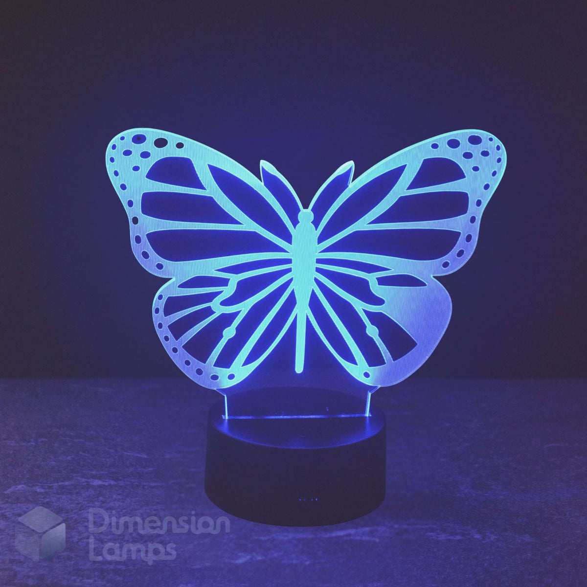 A butterfly-shaped 3D optical illusion night light glowing with a blue light, set on a black cylindrical base against a dark background. A great gift idea for butterfly fans. 
