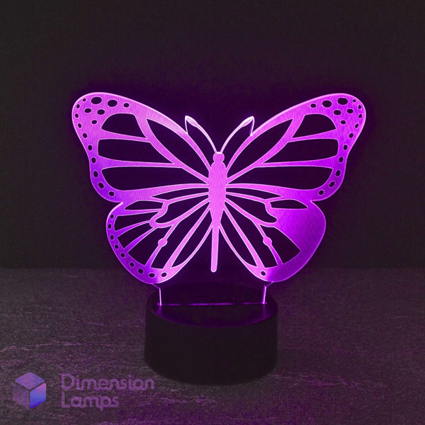 A butterfly-shaped LED night light with intricate cut-out patterns, glowing with a vibrant purple light on a black base. The brand name 'Dimension Lamps' is visible in the bottom-left corner. Butterfly gift lamp.