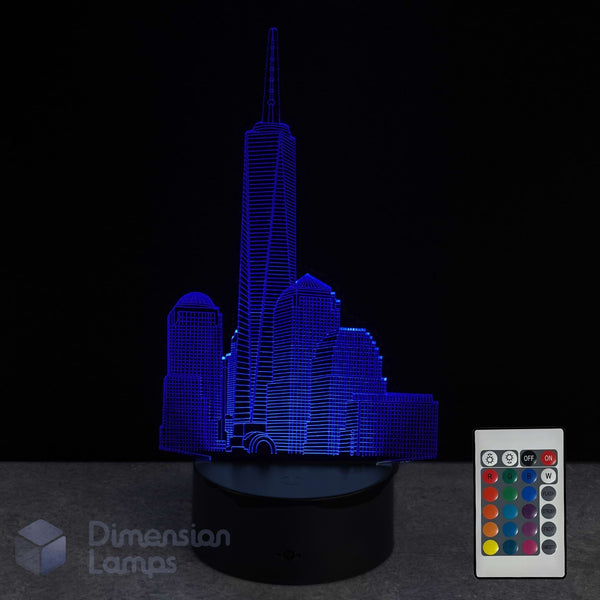 A 3D optical illusion LED night light depicting the One World Trade Center in a city skyline, glowing in vibrant blue on a black base, shown with its white remote control against a black background.