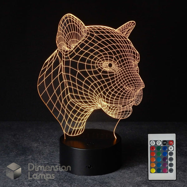 A 3D optical illusion lamp shaped like a panther head, glowing with warm golden light from a black base, shown with a 16 colour remote control on a dark background. The 'Dimension Lamps' logo is visible in the bottom-left corner. Wildlife gift idea. 