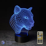 A 3D optical illusion LED night light shaped like a panther or big cat head, glowing with blue light on a black base, shown with a white remote control against a dark background. The Aussie brand name 'Dimension Lamps' is visible as a watermark in the bottom-left corner. Gift idea for Panther lovers.