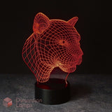 A 3D optical illusion lamp shaped like a panther or big cat head, glowing with red-orange light from thin wire-like lines on a black base. The lamp is photographed against a dark studio background with a grey surface below.