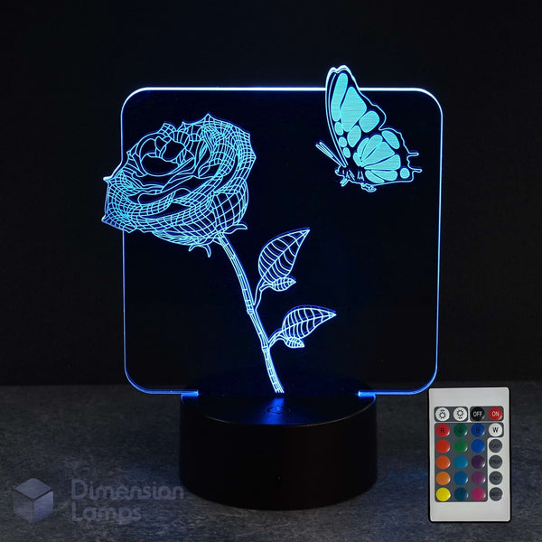 A 3D optical illusion LED night light displaying a glowing blue rose and butterfly on a black base, shown with a remote control against a dark background. A 'Dimension Lamps' watermark is visible in the lower-left corner.