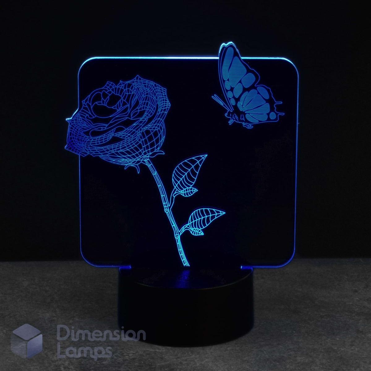 A 3D optical illusion LED night light displaying a glowing blue rose and butterfly design on a black base, set against a dark background.