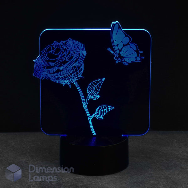 A 3D optical illusion LED night light displaying a glowing blue rose and butterfly design on a black base, set against a dark background.