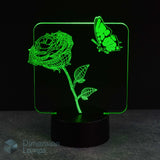 A 3D optical illusion LED night light displaying a glowing green rose and butterfly design on a black base, set against a dark background.