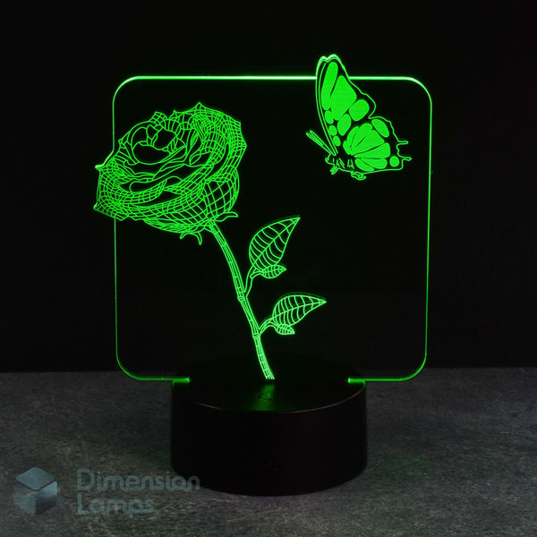 A 3D optical illusion LED night light displaying a glowing green rose and butterfly design on a black base, set against a dark background.