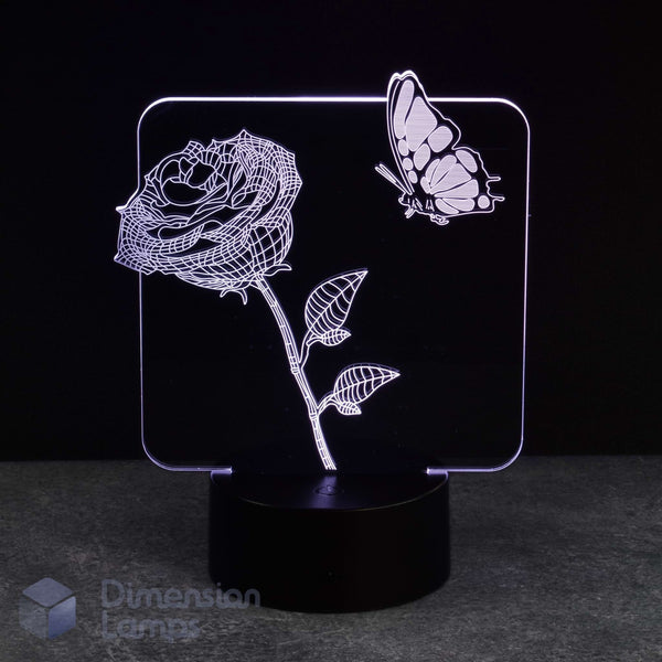 An LED acrylic night light featuring a glowing white line-art rose and butterfly design on a black base, photographed against a dark background.