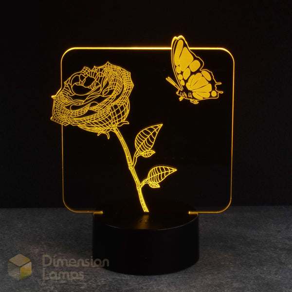 A 3D acrylic LED night light featuring a glowing yellow rose and butterfly design on a black cylindrical base, set against a dark background. A watermark for 'Dimension Lamps' in the bottom-left corner.