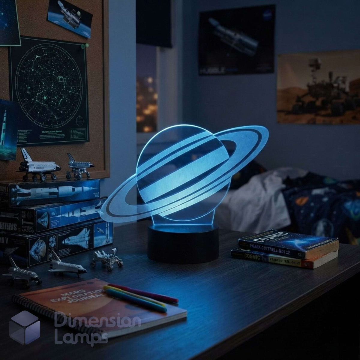 A glowing 3D Saturn night light with a black base on a desk in a bedroom setting, surrounded by books, space models, and posters. Teens space gift.