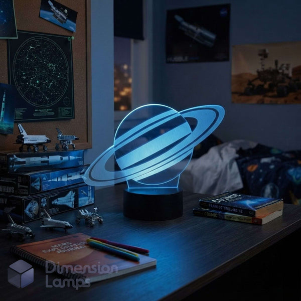 A glowing 3D Saturn night light with a black base on a desk in a bedroom setting, surrounded by books, space models, and posters. Teens space gift.