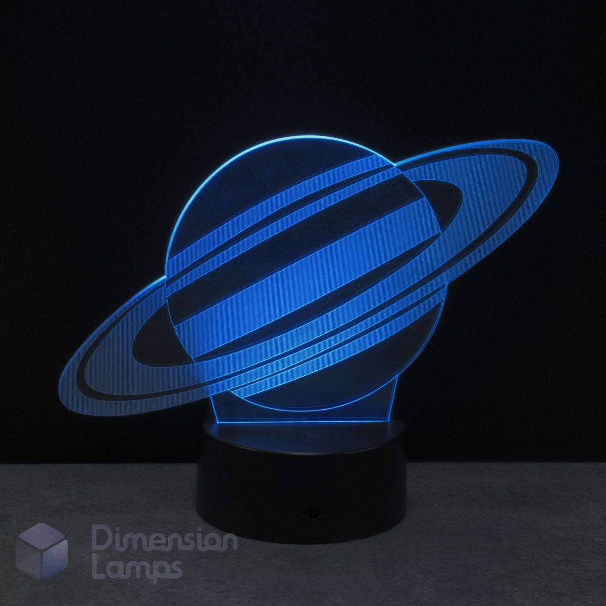 A 3D optical illusion night light of Saturn, glowing bright cyan on a black base against a dark background. Dimension Lamps logo in the bottom corner.