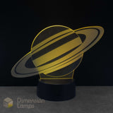 A 3D acrylic illusion lamp shaped like Saturn, glowing with warm golden light, set on a black cylindrical base against a dark background. Space gift.