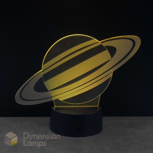 A 3D acrylic illusion lamp shaped like Saturn, glowing with warm golden light, set on a black cylindrical base against a dark background. Space gift.