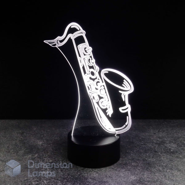 A 3D optical illusion lamp shaped like a saxophone, glowing brightly against a dark background on a black cylindrical base. A 'Dimension Lamps' logo is visible in the bottom-left corner. Saxophonist gift idea.