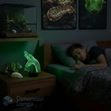 A teenager sleeping in bed in a dark bedroom, with a glowing green 3D-effect lamp shaped like a snake on a bedside table. The room is dimly lit, and framed snake artwork hangs on the wall.