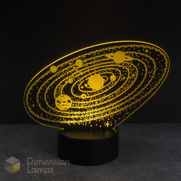 A 3D optical illusion night light shaped like a solar system, featuring glowing yellow planets and orbits on a black acrylic panel mounted on a black cylindrical base. The lamp is photographed against a dark background with a gray surface.
