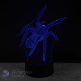 A 3D optical illusion night light shaped like a spider, glowing with blue LED light on a black base against a dark background. A 'Dimension Lamps' logo is visible in the bottom-left corner. Arachnid themed gift idea. 