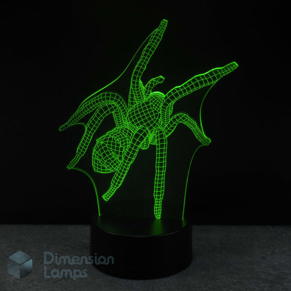 A 3D optical illusion LED night light shaped like a spider, glowing bright green against a black background on a dark base. A 'Dimension Lamps' logo watermark is visible in the bottom-left corner. Arachnid gift idea. 