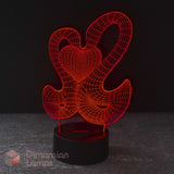 A 3D optical illusion LED night light featuring a 2 interlocking swans forming a red heart on a black cylindrical base, set against a dark background.
