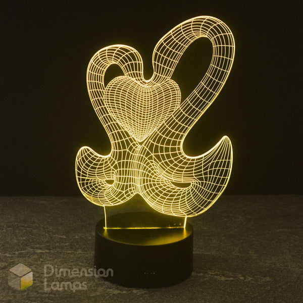 A 3D optical illusion LED night light featuring interlocking swans forming a heart, glowing warm golden light on a black base on a dark background.