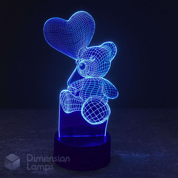 An LED 3D optical illusion LED night light of a teddy bear holding a heart balloon, glowing bright blue on a dark base. Dimension Lamps watermark.