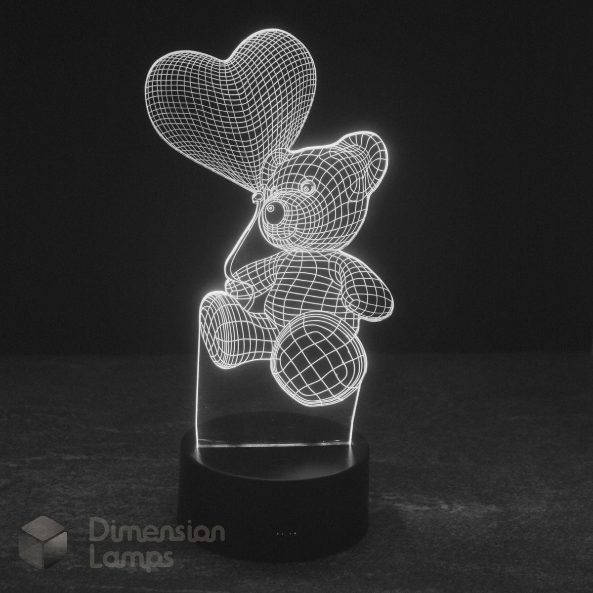 A 3D illusion LED lamp of teddy bear holding a heart balloon glowing white light on a black base against a dark background. Dimension Lamps watermark.