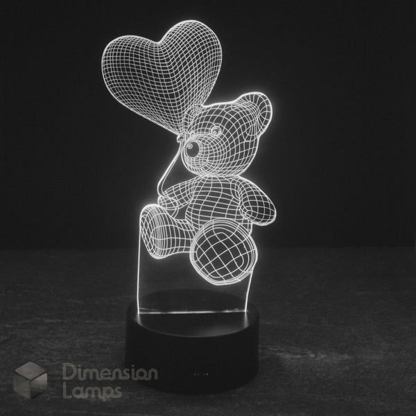 A 3D illusion LED lamp of teddy bear holding a heart balloon glowing white light on a black base against a dark background. Dimension Lamps watermark.