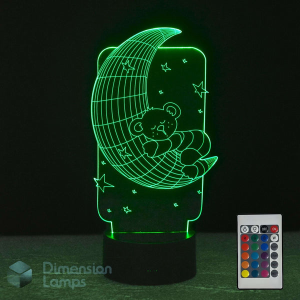 A 3D optical illusion night light of a teddy bear sleeping on a crescent moon, glowing green in the dark. A black base and a 16 colour remote control.