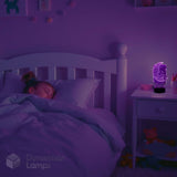 A 3D illusion night light shaped like a cartoon character glows purple on a bedside table next to a bed where a child is sleeping, by Dimension Lamps.