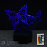 A 3D optical illusion LED night light with two butterflies on a black base, shown with a 16 colour remote control against a dark background. 
