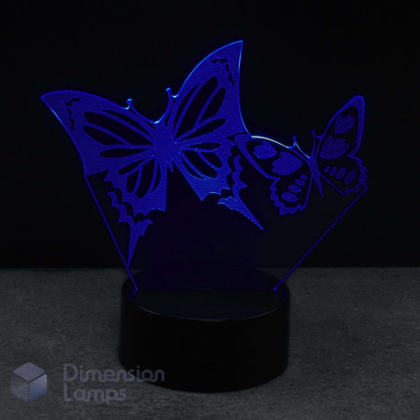 A 3D acrylic illusion lamp featuring two glowing blue butterflies on a black base against a dark background with a 'Dimension Lamps' logo.
