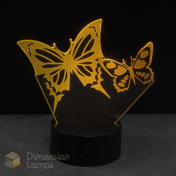 A 3D optical illusion night light featuring two glowing golden-yellow laser-cut butterflies on a black base, set against a dark background.